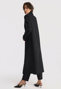 Czarny wełniany płaszcz o dopasowanym kroju, długiej sylwetce i kołnierzu w kształcie litery V. Zestawiony z czarnymi spodniami i butami na obcasie ze zapięciem.