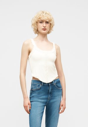 Persona rubia con cabello rizado que lleva una blusa sin mangas blanca con volantes y jeans azules, posando contra un fondo liso y claro.