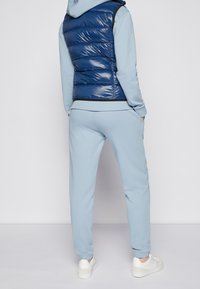 Person som bär ljusblå sweatpants och en matchande huvtröja under en skinande blå ärmlös pufferväst, står med händerna i fickorna.