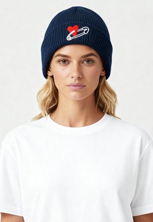 Blauwe ribgebreide muts met een rood hart en een witte veiligheidsspeld op de voorkant. Model draagt een effen wit T-shirt.