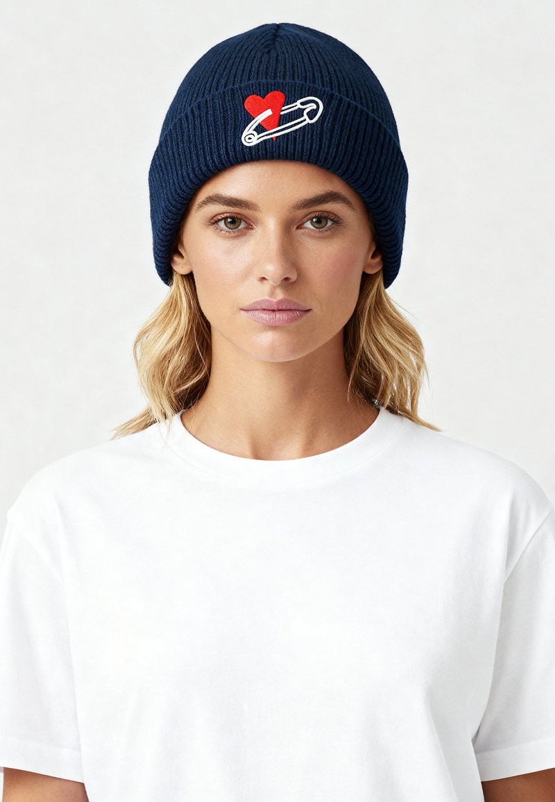 Cappello a maglia blu a coste con un design sul davanti di un cuore rosso e una spilla da balia bianca. Il modello indossa una maglietta bianca semplice.