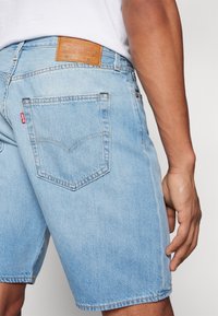 Mies, joka on pukeutunut vaaleansinisiin Levi's-farkkushortseihin ja valkoiseen paitaan, näkyy takaa vyötäröstä puoliväliin reittä, takatasku ja etiketti näkyvissä.