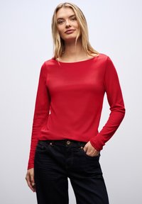 Rode lange mouwen top van gladde stof met een brede halslijn, gecombineerd met donkerblauwe jeans met zichtbare stiksels en zakdetails.