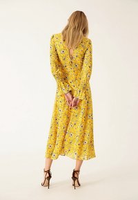 Femme aux longs cheveux blonds, debout, les mains derrière le dos, portant une robe midi jaune à fleurs et des sandales marron à talons hauts.