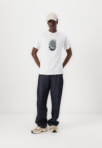 White cotton t-shirt with a colorful graphic design on the chest, paired with dark denim pants and beige athletic shoes. Light-colored cap.