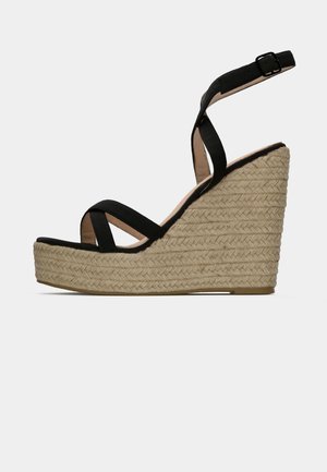Schwarze Keil-Sandalen mit Kreuzriemen-Design, strukturiertem Jute-Plateau und verstellbarem Knöchelriemen. Die Seitenansicht betont ihre Höhe und Form.