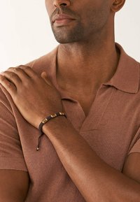 Fläkt brun armband med gyllene geometriska detaljer, bärs på handleden mot en brun polo. Slät textur, justerbar snörstängning.