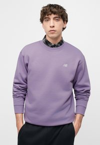 Νεαρός με κατσαρά μαλλιά που φοράει μοβ μπλούζα με στρογγυλή λαιμόκοψη πάνω από ένα πουκάμισο με ασπρόμαυρο καρό, τα χέρια στις τσέπες.