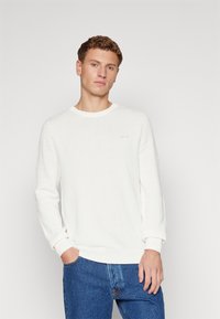Witte langharige trui met een textuur gebreide ontwerp, ronde hals, geribbelde manchetten en onderrand. Kleine branding zichtbaar op de borst. Gecombineerd met blauwe jeans.