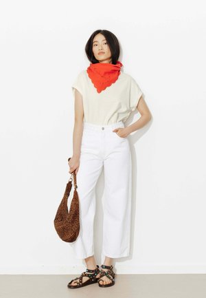 Femme debout contre un mur blanc, portant un pantalon blanc, une chemise crème, un foulard rouge autour du cou, tenant un sac imprimé léopard avec des sandales noires à clous.