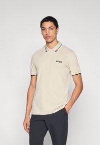 Bézs színű, puha anyagból készült galléros póló, fekete-fehér csíkos gallérral, rövid ujjakkal és a bal mellrészen "BOSS" logóval.