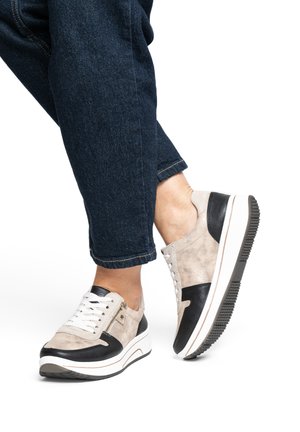 Person trägt beige-schwarze Sneakers mit weißen Schnürsenkeln und Reißverschluss-Details, kombiniert mit dunkelblauen hochgekrempelten Jeans, vor einem weißen Hintergrund.