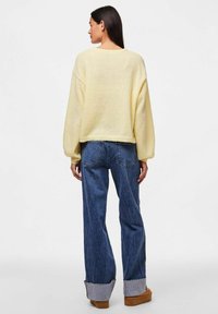 Rumena pletena jopica z širokimi rokavi, v kombinaciji z modrimi hlačami z visokim pasom in zloženim robom. Mehka tekstura in sproščen kroj.