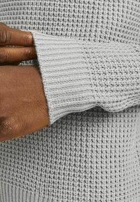Suéter de punto gris, con un patrón de puntadas abiertas texturizadas y puños acanalados. El material parece suave con un acabado ligeramente elástico.