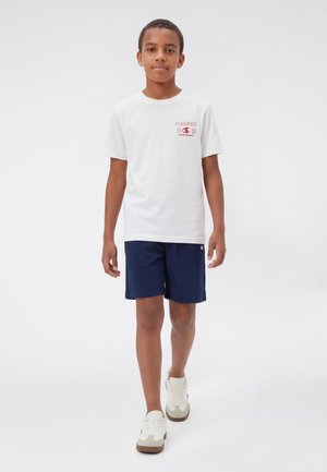 Junge, der in Richtung Kamera geht, trägt ein weißes Champion-T-Shirt, marineblaue Shorts und weiße Sneakers vor weißem Hintergrund.