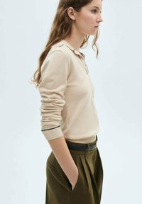 Pull beige avec col, manches longues et poignets côtelés. Associé à un pantalon taille haute vert olive et une ceinture noire.