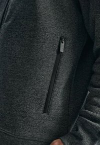 Szara polarowa kurtka zamykana na zamek, z zamykaną na zamek boczną kieszenią i ściągaczami na rękawach, o gładkiej fakturze i subtelnym melanżowym wzorze.