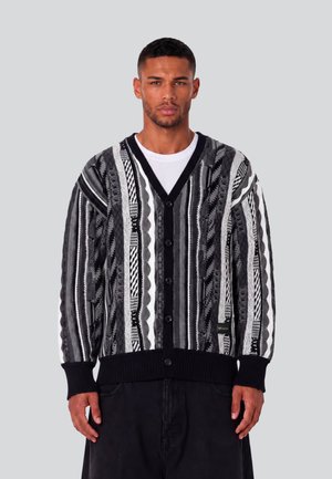Mand iført en sort, hvid og grå mønstret strikket cardigan over en hvid skjorte, stående mod en ensartet lys grå baggrund.