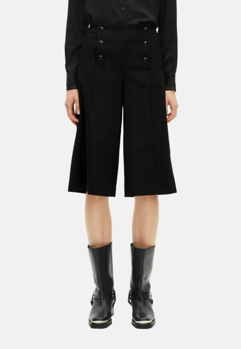 Culotte neri a vita alta con due file di bottoni sul davanti e design plissettato, abbinati a stivali in pelle nera alla caviglia.