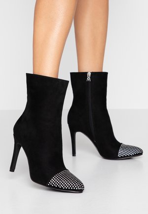 Stivaletti in suede neri alla caviglia con punta a punta, decorati con una punta borchiata e una zip laterale per la chiusura. Il tacco a stiletto aumenta l'altezza.