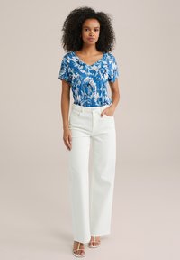 Zila ziedu īsām piedurknēm augšdaļa ar V-veida kakla izgriezumu; kombinēta ar baltām platām biksēm un baltām sandālēm ar siksniņām; gluda auduma un brīvs piegriezums.