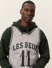 Hallhallane võrgusilma korvpallimüts, millel on mustade tähtedega trükitud "LES DEUX" ja "11". Seljas tume rohelise kapuutsiga püksi, millel on nööriga kapuuts.