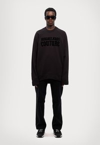 Prevelik crni dukserica s logotipom "VERSACE JEANS COUTURE", u kombinaciji s tamnim trapericama i crnim tenisicama, nosi model s naočalama za sunce.