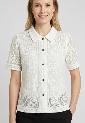 Frau trägt eine weiße, kurzärmelige Spitzenbluse mit Blumenmustern und braunen Knöpfen, steht mit einer Hand in der Tasche vor einem einfarbigen Hintergrund.
