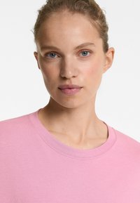 Tricou scurt cu mânecă scurtă, de culoare roz deschis, fabricat din bumbac moale. Prezintă un decolteu rotund și un design simplu cu detalii subtile de cusătură.