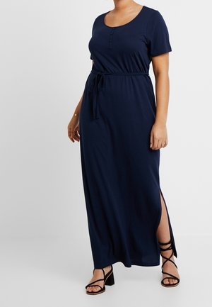 Femme portant une robe maxi bleu marine à manches courtes avec ceinture à la taille, fente latérale, et sandales noires à talons blocs à brides.