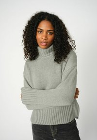 Lichtgrijze turtleneck trui met geribbelde manchetten en zoom, voorzien van een geanimeerd breipatroon. Gecombineerd met donkere jeans.