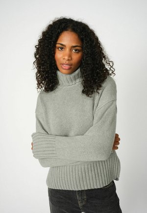 Lichtgrijze turtleneck trui met geribbelde manchetten en zoom, voorzien van een geanimeerd breipatroon. Gecombineerd met donkere jeans.