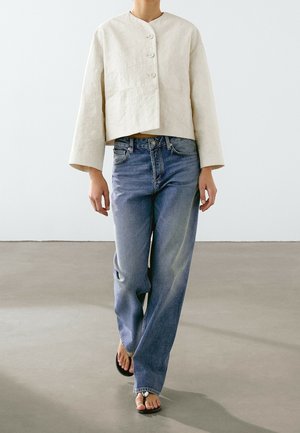 Personne marchant vêtue d'une veste crème texturée boutonnée, d'un jean bleu ample et de tongs noires sur un sol gris lisse.