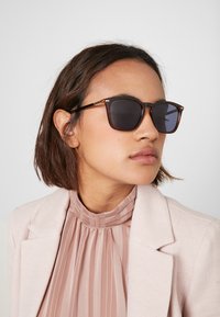 Bruna sköldpaddsrektangulära solglasögon med mörka linser, gyllene detaljer och en elegant design. Bärs med en plisserad ljusrosa blus och kavaj.