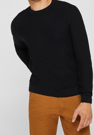 Mann trägt einen schwarzen Pullover mit langen Ärmeln und senffarbene Hose, steht vor einem neutralen Hintergrund.