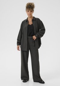 Donna con capelli ricci che indossa un completo a righe grigio scuro con camicia morbida e pantaloni larghi, top in pizzo nero e scarpe nere, in piedi con sicurezza.