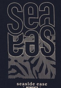 Tessuto blu navy con testo ricamato "sea gos" sopra un motivo di onda e sole e la scritta "seaside ease moments" stampata sotto in beige chiaro.