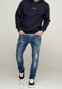 Sudadera navy con bolsillo frontal, combinada con jeans azules desgastados con rotos. Llevada con zapatillas negras y blancas, mostrando un look casual.