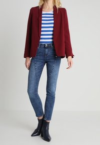 Kastanienbrauner maßgeschneiderter Blazer über einem blau-weiß gestreiften Oberteil, kombiniert mit engen Denim-Jeans und schwarzen Stiefeletten. Glatter Stoff, moderner Schnitt.