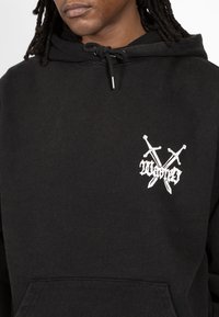 Musta huppari, joka on valmistettu pehmeästä kankaasta, jossa on edessä kengurutasku, säädettävä kiristysnauhallinen huppu ja valkoinen ristikkäin miekkoja esittävä grafiikka.