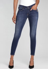 Jeans ajustados en denim azul oscuro, con un ligero desgastado, diseño de cinco bolsillos, cierre frontal con botón y lazo para cinturón estándar.