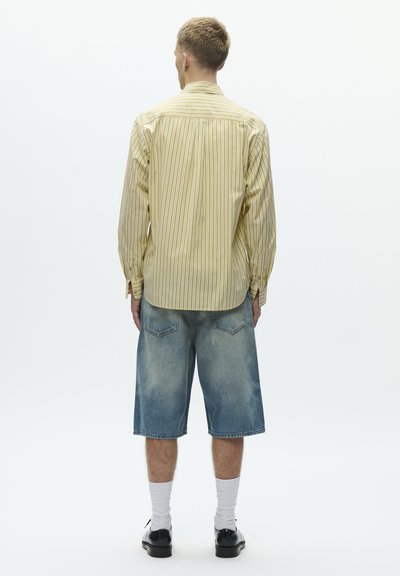 Keltainen raidallinen pitkähiha-paita, yhdistettynä löysiin farkkushortseihin. Lyhyissä housuissa on metalliset napit ja kaksi takataskua.