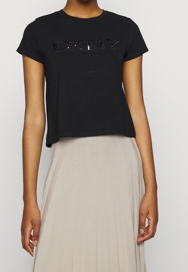 T-shirt crop nero in cotone morbido, con il logo "DKNY" in paillettes nere lucide sul davanti. Abbinato a una gonna plissettata beige chiaro.