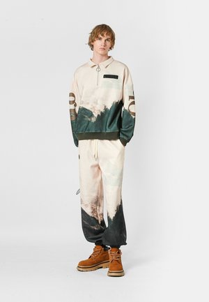 Suéter claro con media cremallera y un estampado gráfico en verde y beige; los joggers a juego presentan un diseño similar, combinados con botas marrones.