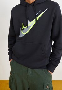 Sudadera negra con un gran logo de Nike en gris y verde neón. Presenta cordones ajustables y un bolsillo frontal. Tejido texturizado, corte casual.