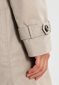 Dłoń oparta o rękaw beżowego trencza z przeszytym paskiem i detalem z tortoishell guzika.