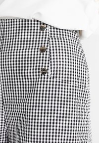 Černobílá kostkovaná sukně s vysokým pasem, která má tři kovové knoflíky po straně a texturovaný povrch.