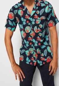 Lyhythihainen paita tummansinistä kangasta, jossa on trooppinen hedelmä- ja kukkakuosi kirkkaissa väreissä, edessä napillinen ja kaulus.