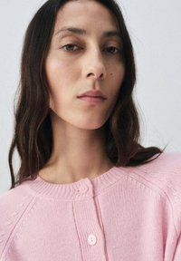 Gilet en tricot rose avec un col rond, une fermeture à boutons et un motif texturé sur les manches. Tissu doux avec une finition lisse.