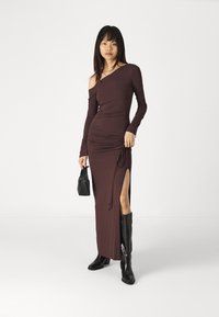 Braun, schulterfreies Kleid mit langen Ärmeln, geraffter Textur, seitlichem Schlitz und Kordelzugdetails, kombiniert mit schwarzen Stiefeletten und einer Handtasche.